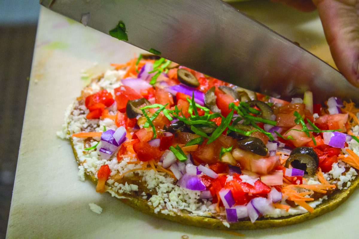 Brian cuts a raw pizza with onion bread, tomato sauce, cauliflower to act as the cheese, carrots, pappers, onions, olives, tomatoes, and basil sauce.